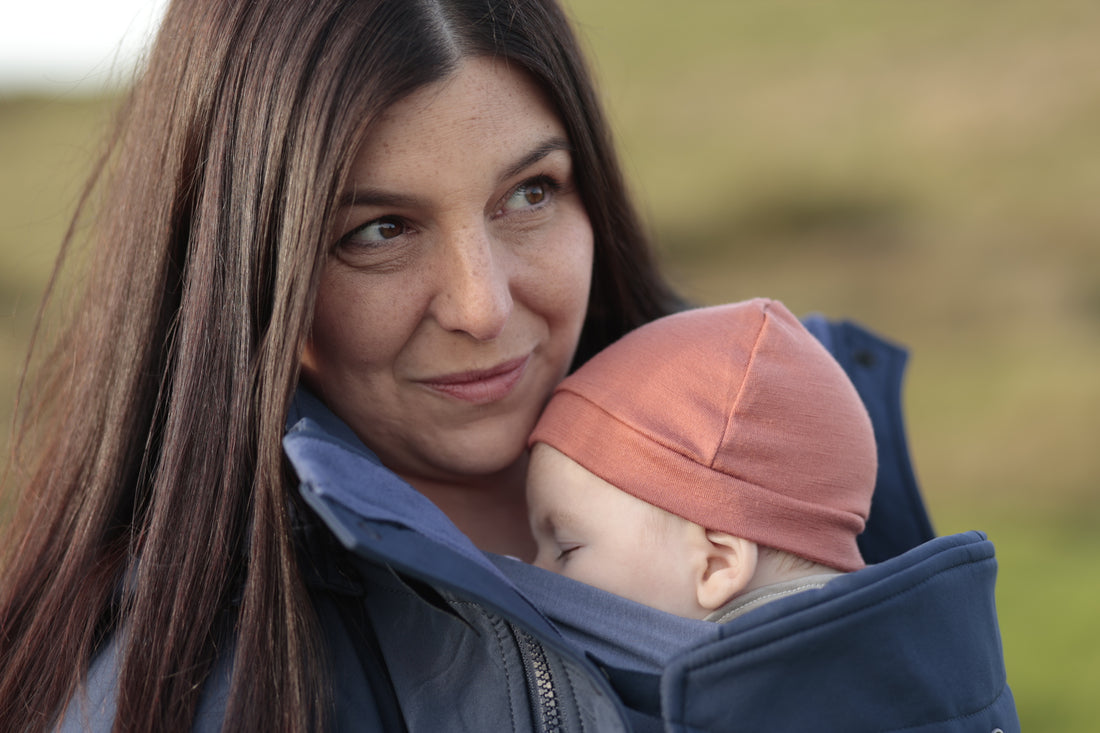 Baby wearing soft pink wool hat snuggled into mother's chest inside her sling and babywearing coat while outdoors