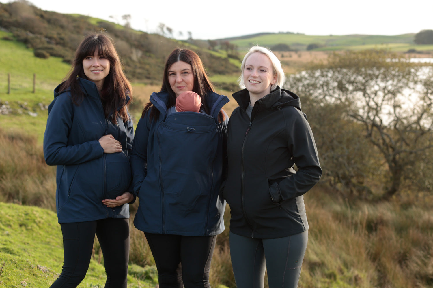 Three women standing together on a hillside with a lake behind. One is pregnant and cupping her bump, and one woman has a baby zipped into her babywearing jacket