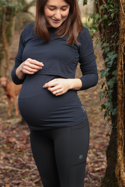 Pregnant woman standing in woodland looking down and smiling, wearing navy long sleeve top and black maternity leggings